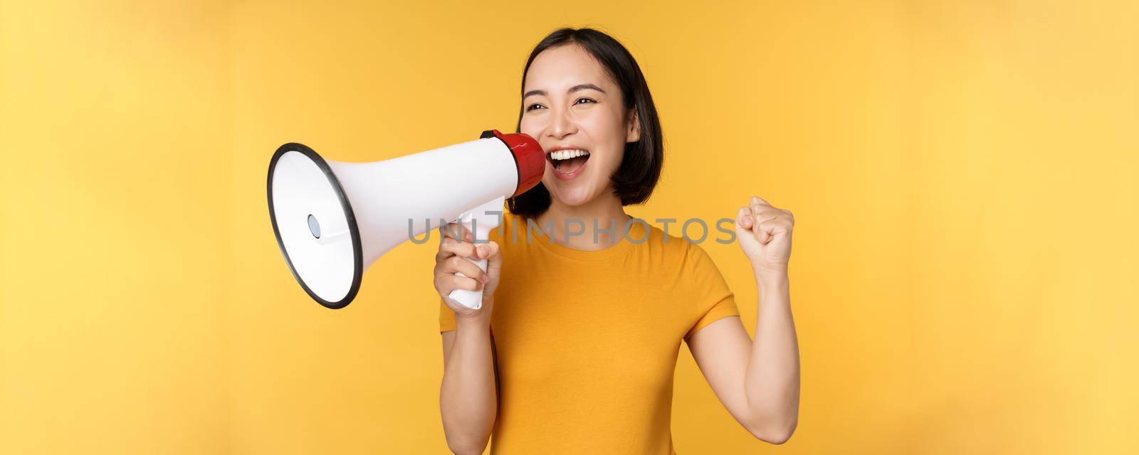 Announcement. Happy asian woman shouting loud at megaphone, recruiting, protesting with speaker in hands, standing over yellow background.