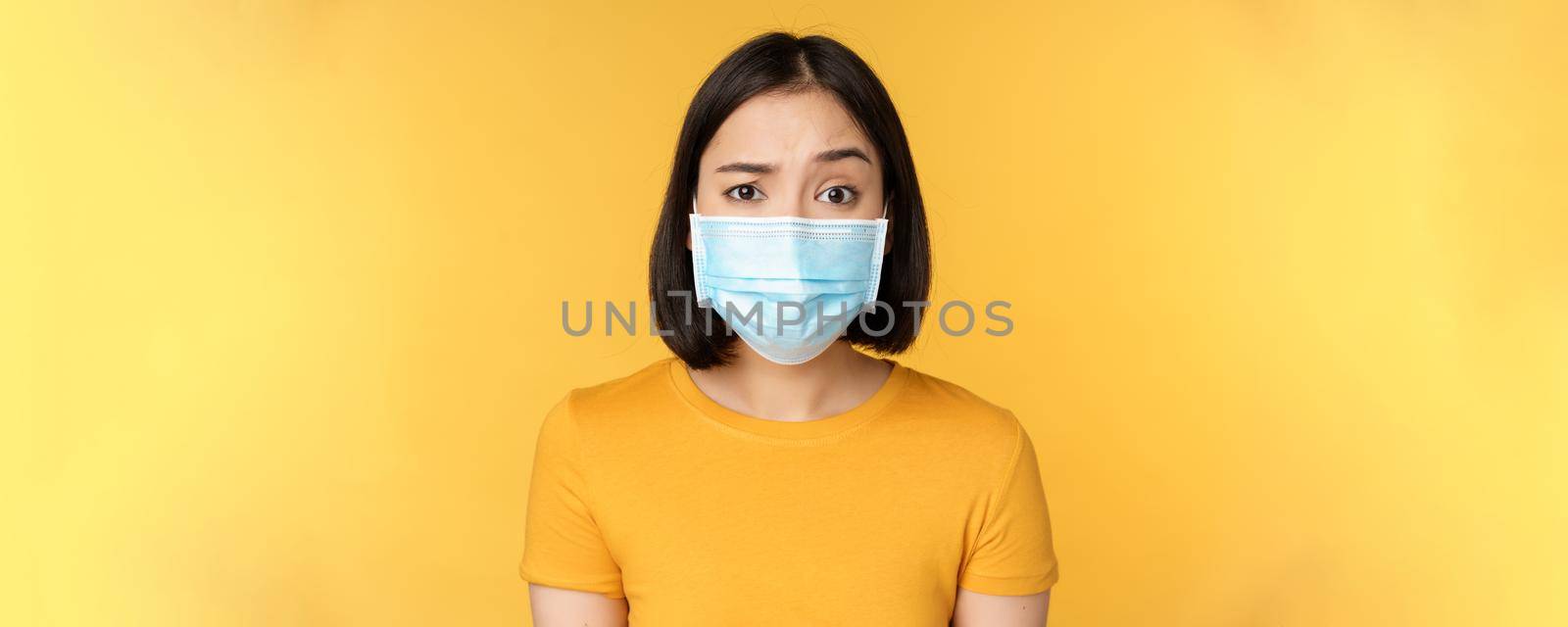Portrait of skeptical and confused asian woman in medical face mask, raising eyebrow doubtful, standing over yellow background by Benzoix