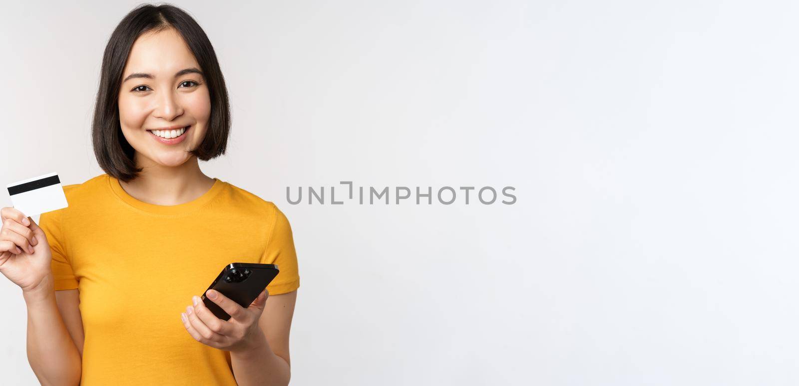 Beautiful smiling asian girl using credit card and mobile phone, paying online on smartphone, standing in yellow tshirt over white background.