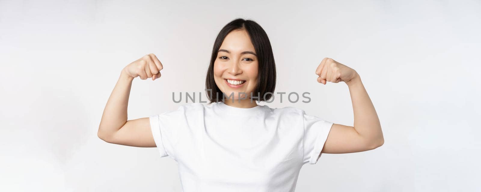Smiling asian woman showing flexing biceps, muscles strong arms gesture, standing in white tshirt over white background by Benzoix