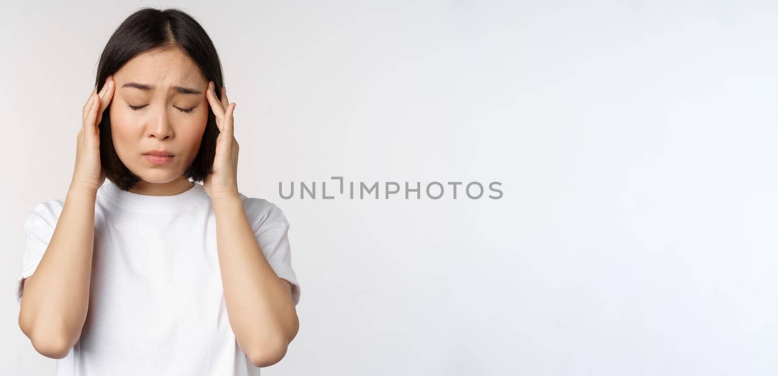 Portrait of asian girl feeling headache, migraine or being ill, standing in white t-shirt over white background. Copy space