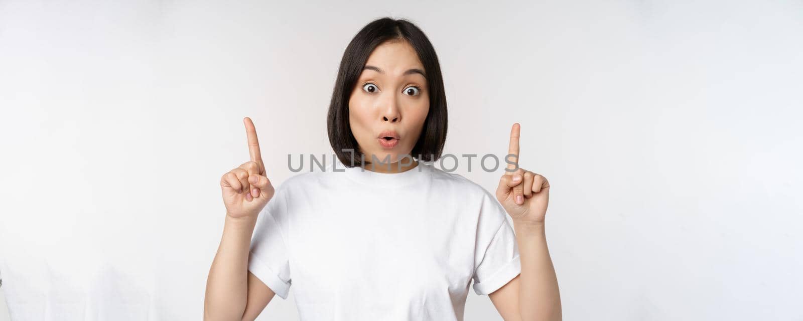 Portrait of enthusiastic young woman, asian girl smiling pointing fingers up, showing advertisement upwards, standing over white background.