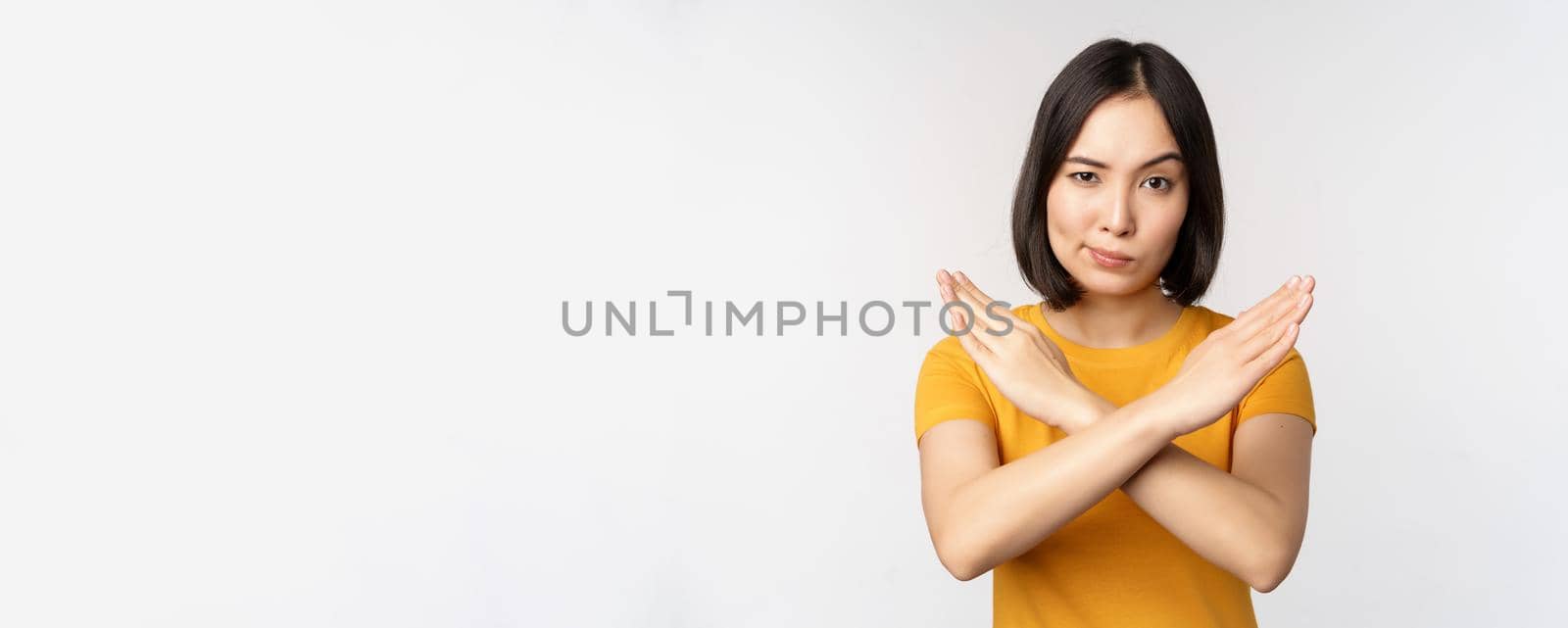 Portrait of asian woman looking serious and angry, showing stop prohibit gesture, taboo sign, forbidding smth, standing in yellow tshirt over white background.