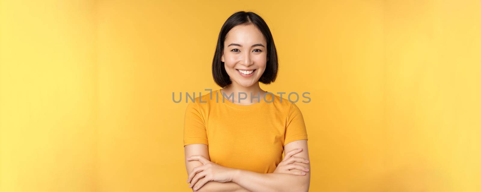 Confident asian girl cross arms on chest, smiling and looking assertive, standing over yellow background by Benzoix