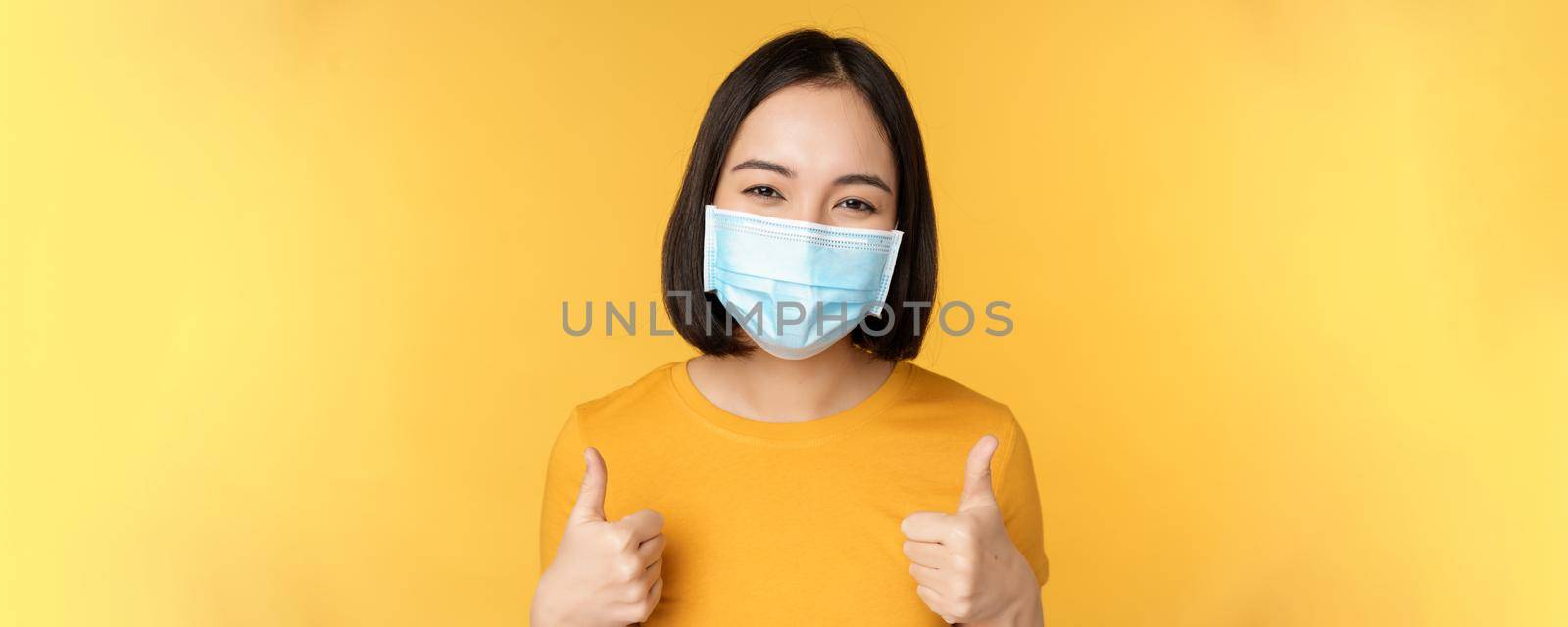 Cheerful korean woman in medical face mask, support people during pandemic, wear personal protective equipment from covid-19, showing thumbs up in approval, yellow background.