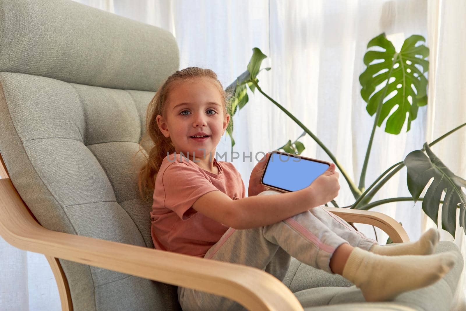 Side view of adorable little girl with long blond hair in casual clothes smiling and looking at camera while sitting on comfortable armchair and demonstrating smartphone with blank screen