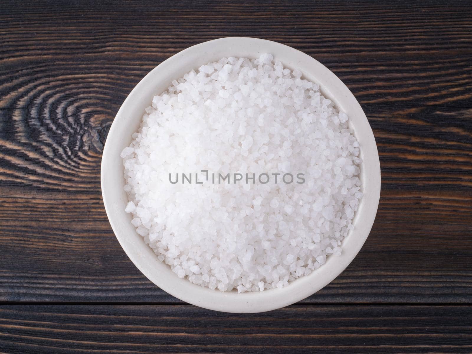White bowl with large sea salt on brown bright dark wooden table. Top view.