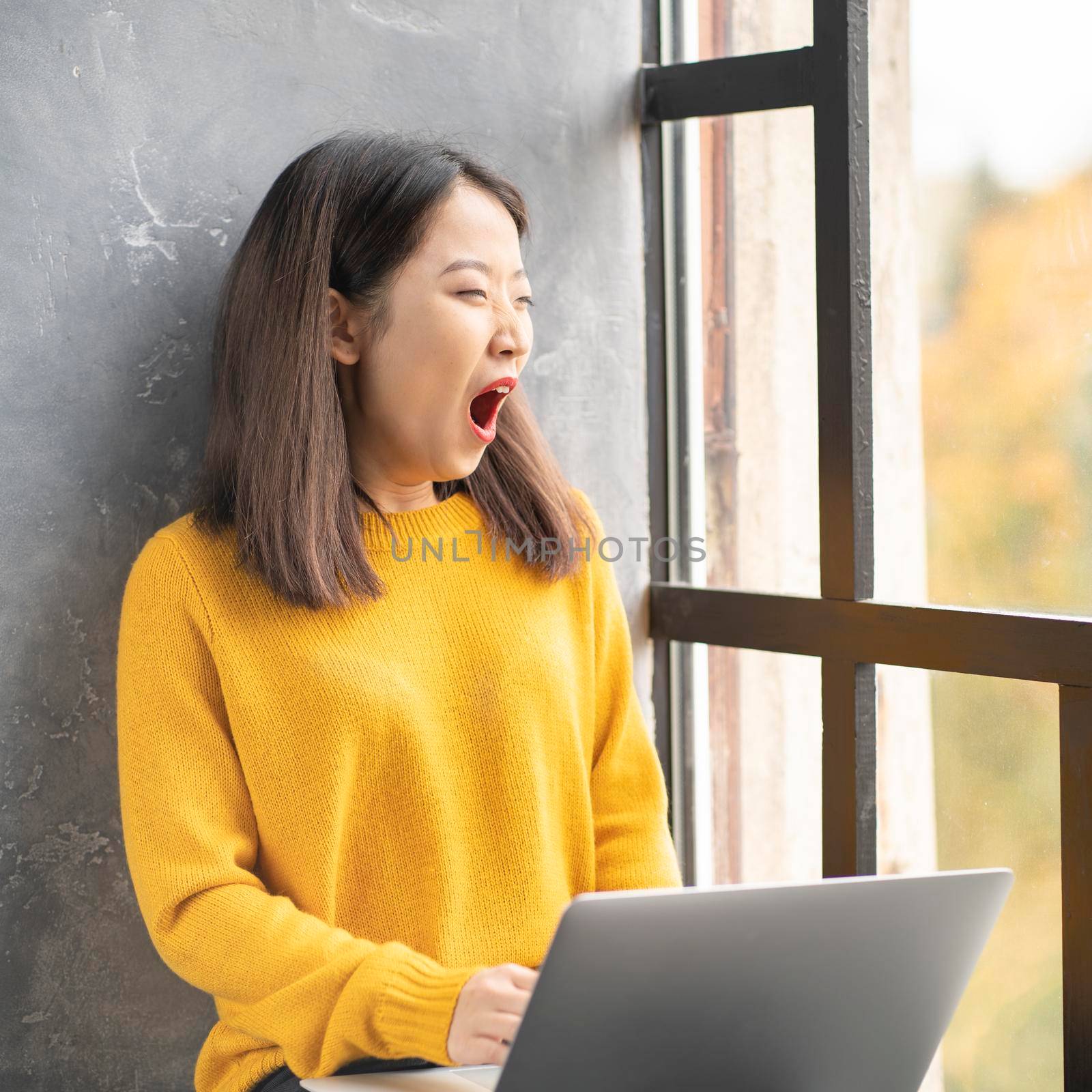 Asian woman yawning due to overworking and exhausting. Young lady in bright yellow jumper working on laptop at home or in cafe. Business oriental female