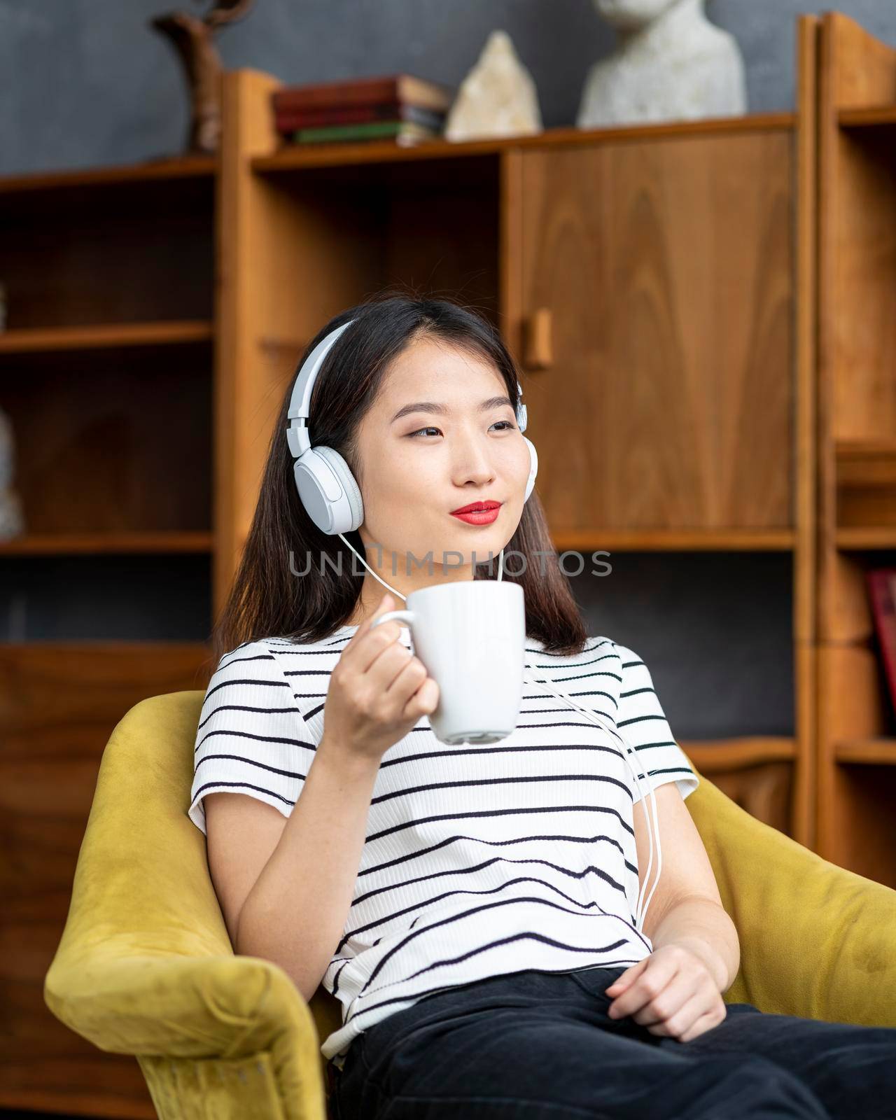 Young beautiful Asian girl listening to music with headphones sitting in chair and looking out window. Woman drinking coffee from mug. Morning routine and contemplation