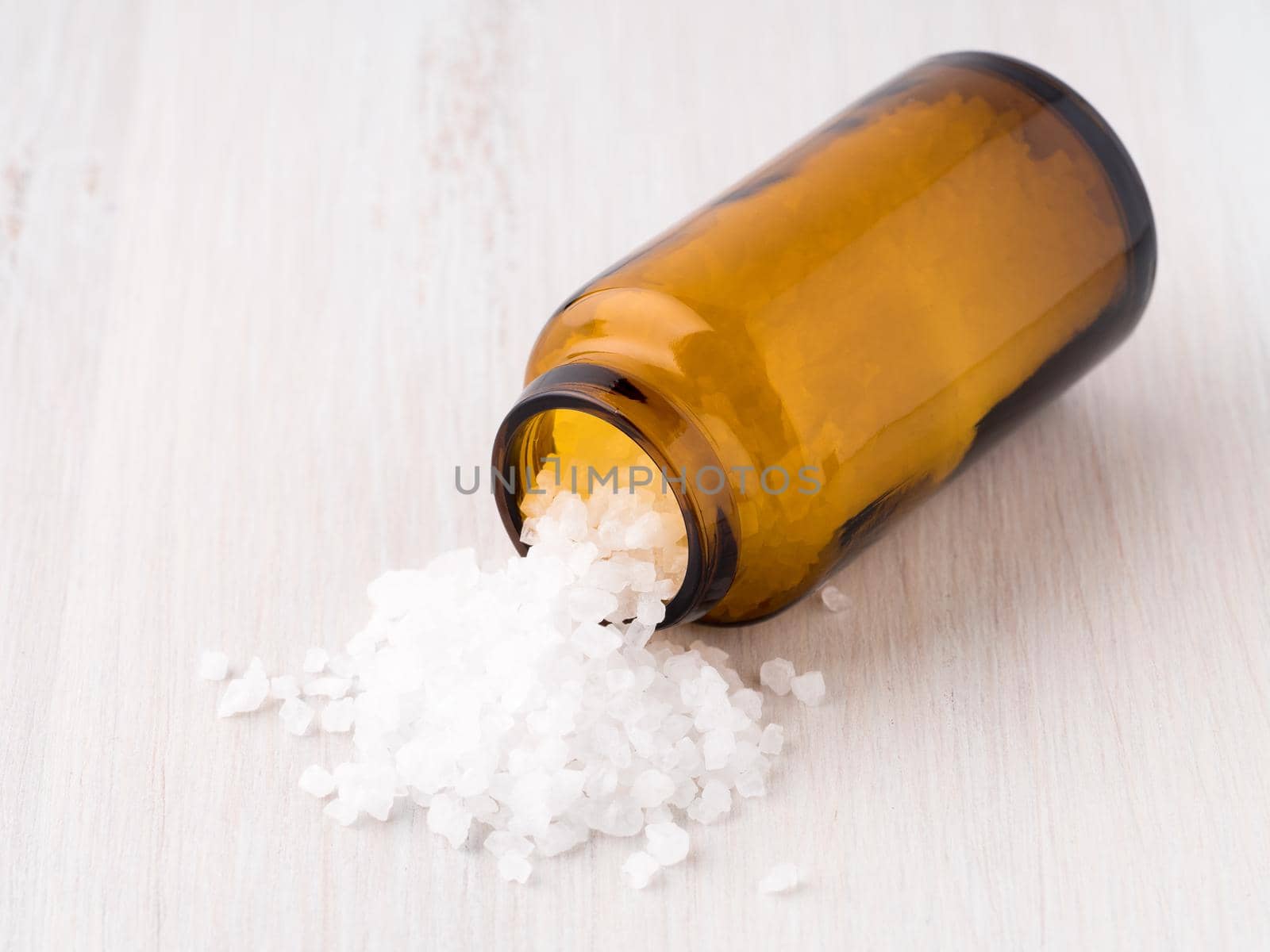 glass jar with large sea salt, lies on a light wooden table, scattered salt crystals.