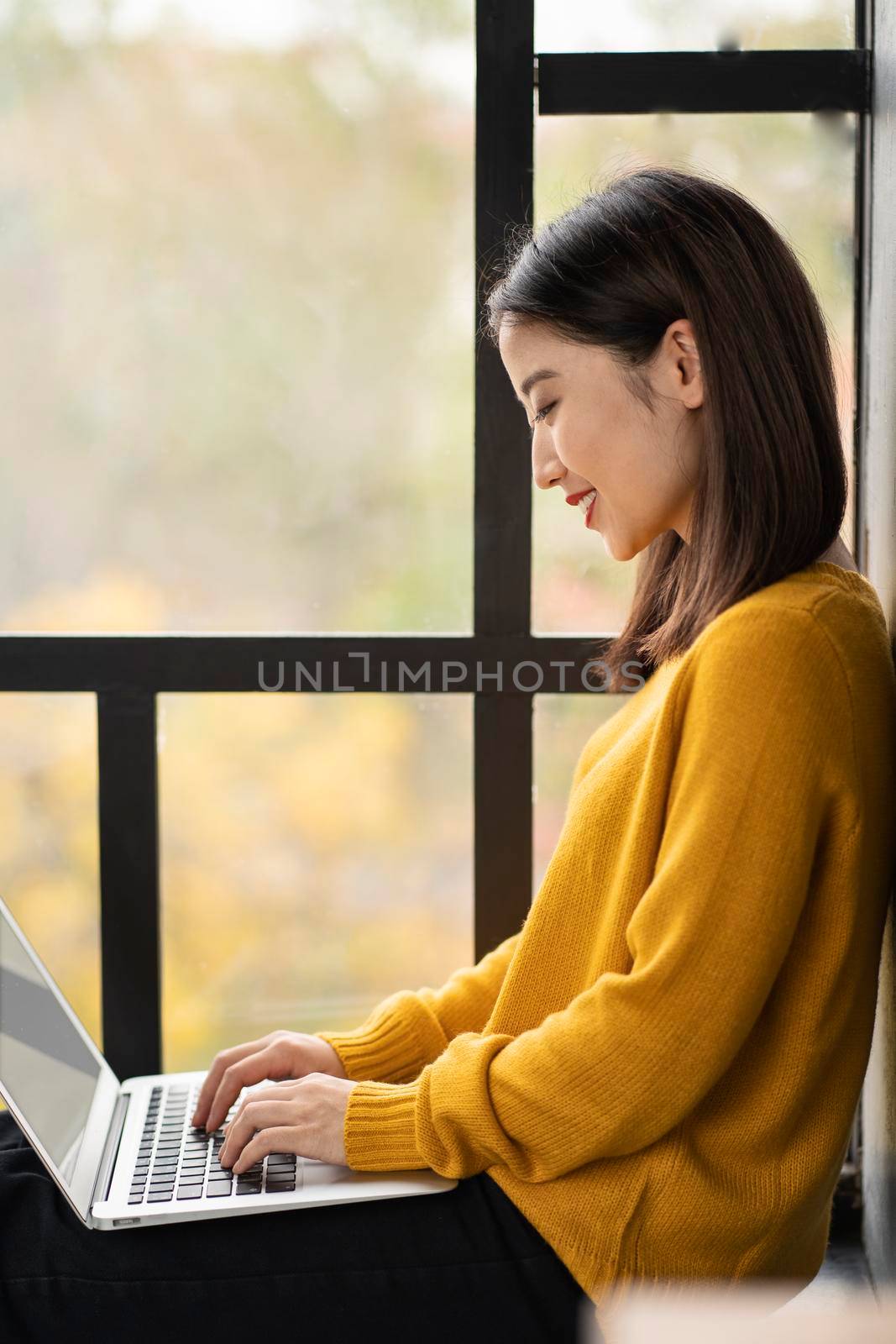 Asian woman working on laptop at home or in cafe on window. Young female in bright yellow jumper is sitting and typing on computer. Business oriental lady in windowsill