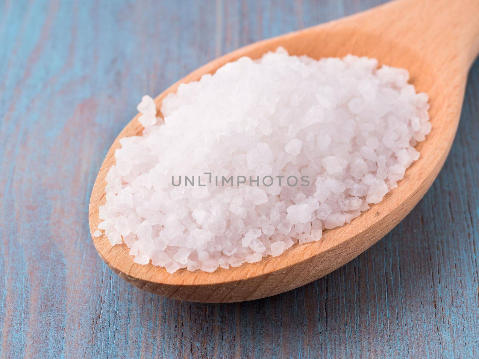large wooden spoon with big sea salt crystals on the bright blue wooden background, side view