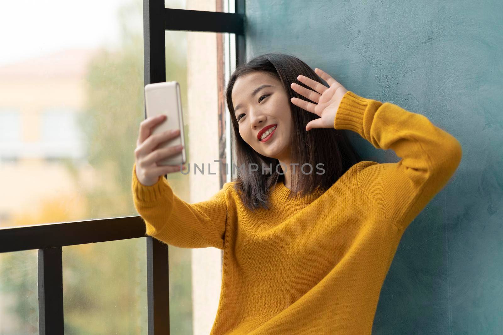 Young Asian woman takeing selfie and waving hand. Beautiful teenage girl with bright red lips siting on window and communicating via video call with boyfriend