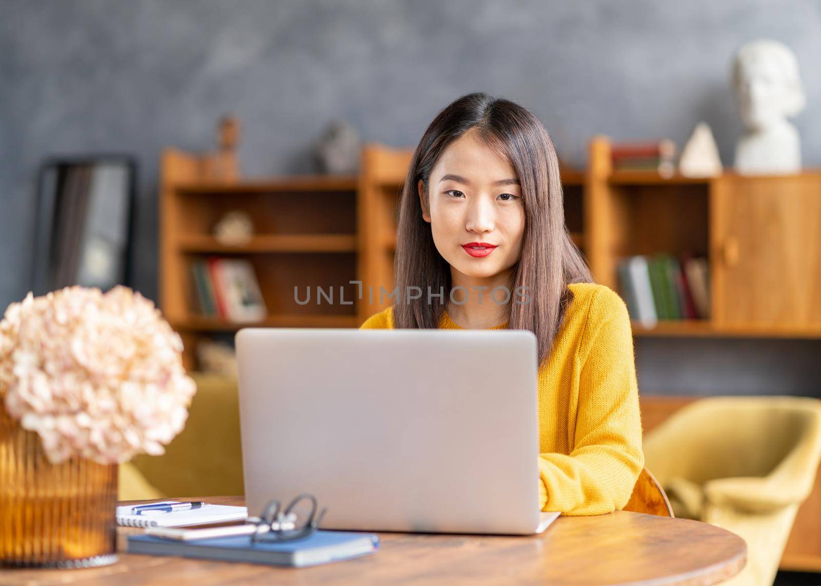 Asian woman working on laptop at home or in cafe. Young lady in bright yellow jumper by NataBene