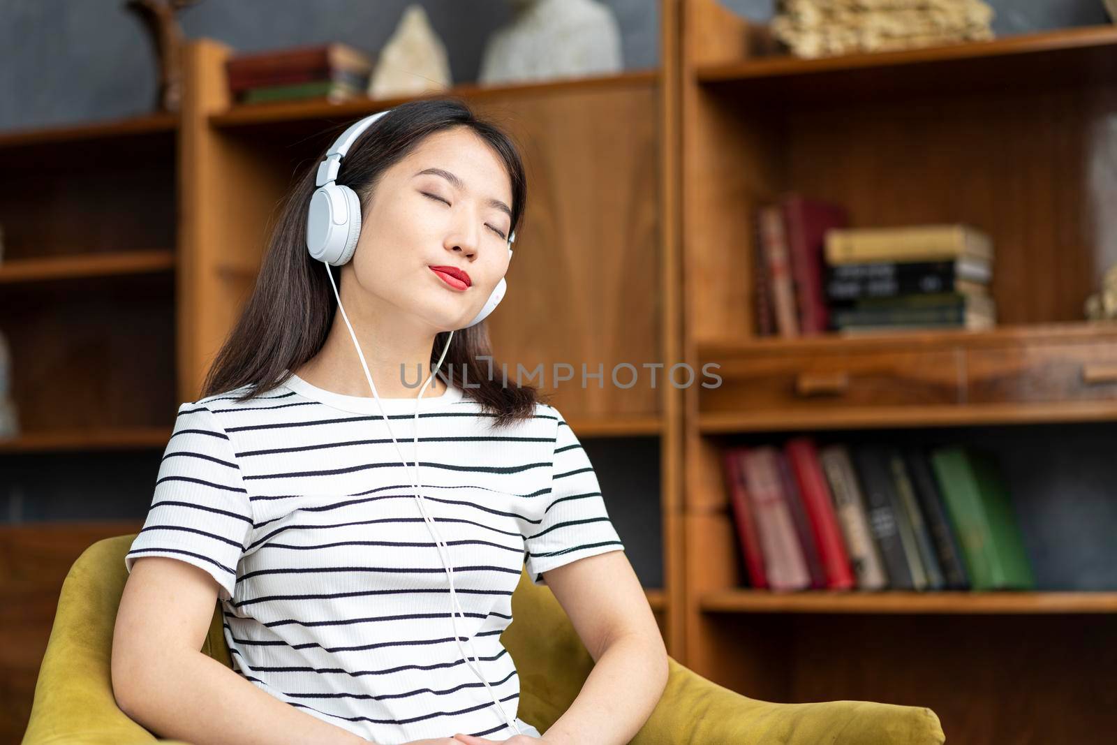 Young beautiful Asian girl listening to music with headphones sitting in chair. Woman with closed eyes. Morning routine and contemplation