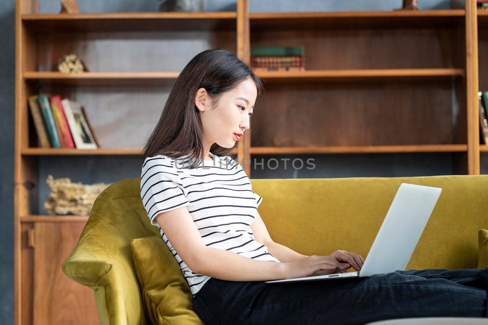 Asian woman looking at laptop, working at home, liying on sofa. Beautiful Japanese female freelancer smiling and typing at computer in hotel on couch