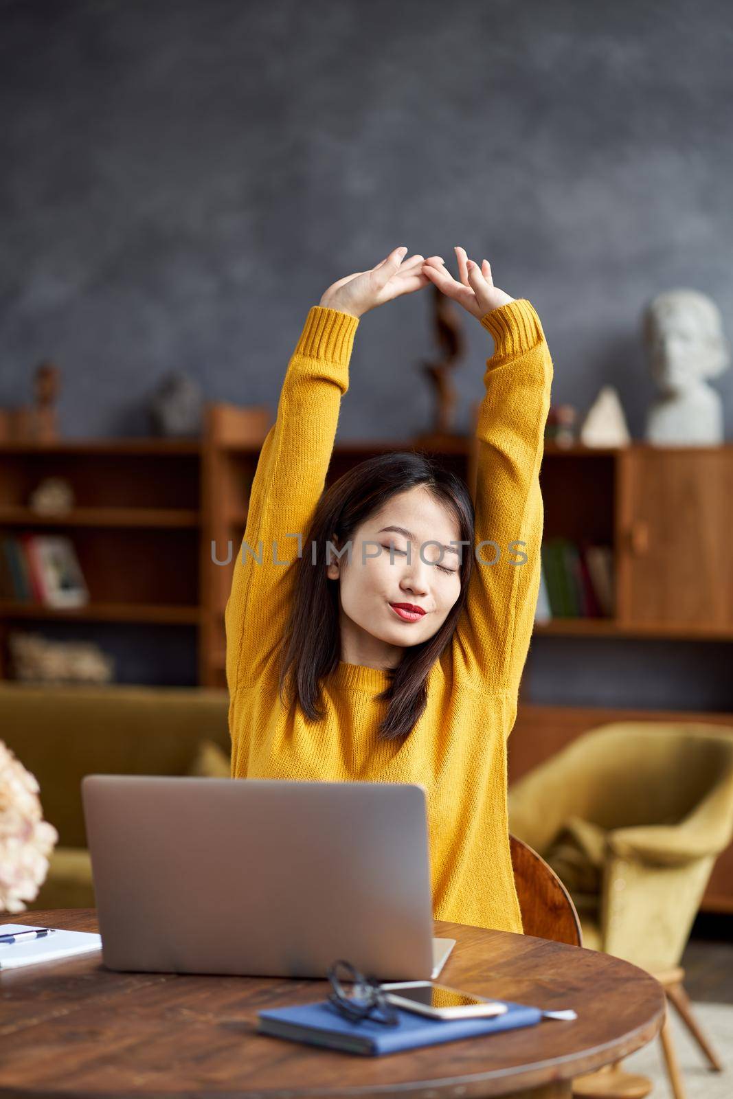 Asian woman working on laptop at home or in cafe stretching tiredly, pulling her hands up. Young lady in bright yellow jumper sits at desk. Vertical