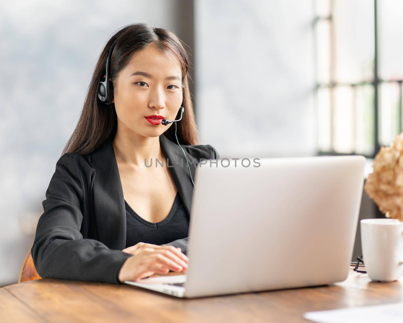 Happy asian businesswoman in headset speaking by conference call and video chat on laptop in office, smiling chinese support service agent consulting customer. Female portrait looking at camera