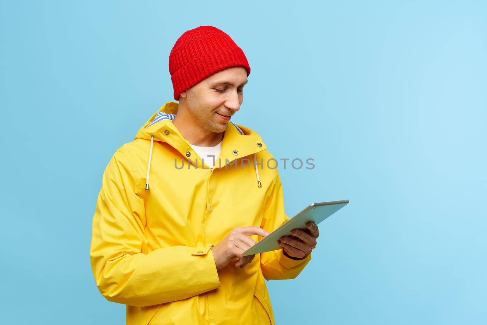 Portrait of positive cool man in warm autumn top outdoor clothes holding tablet, male isolated over blue color background. Bright casual wear, yellow cloak