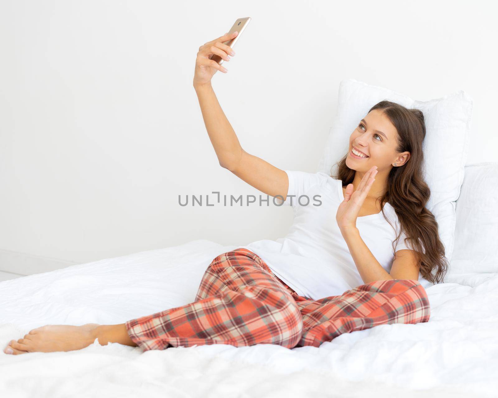 Pretty female selfie, blonde takes photo on mobile phone laying in bed and waving hand by NataBene