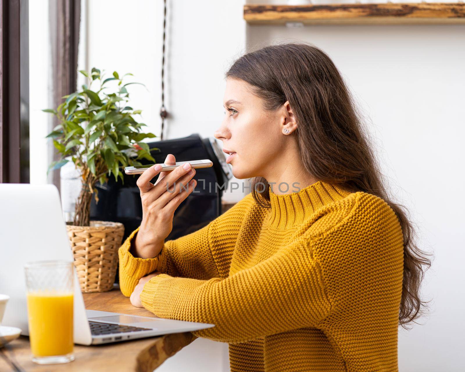 Portrait of positive young woman recording audio message, speaking to microphone of mobile phone.Female on bright yellow jumper. Work from cafe or home