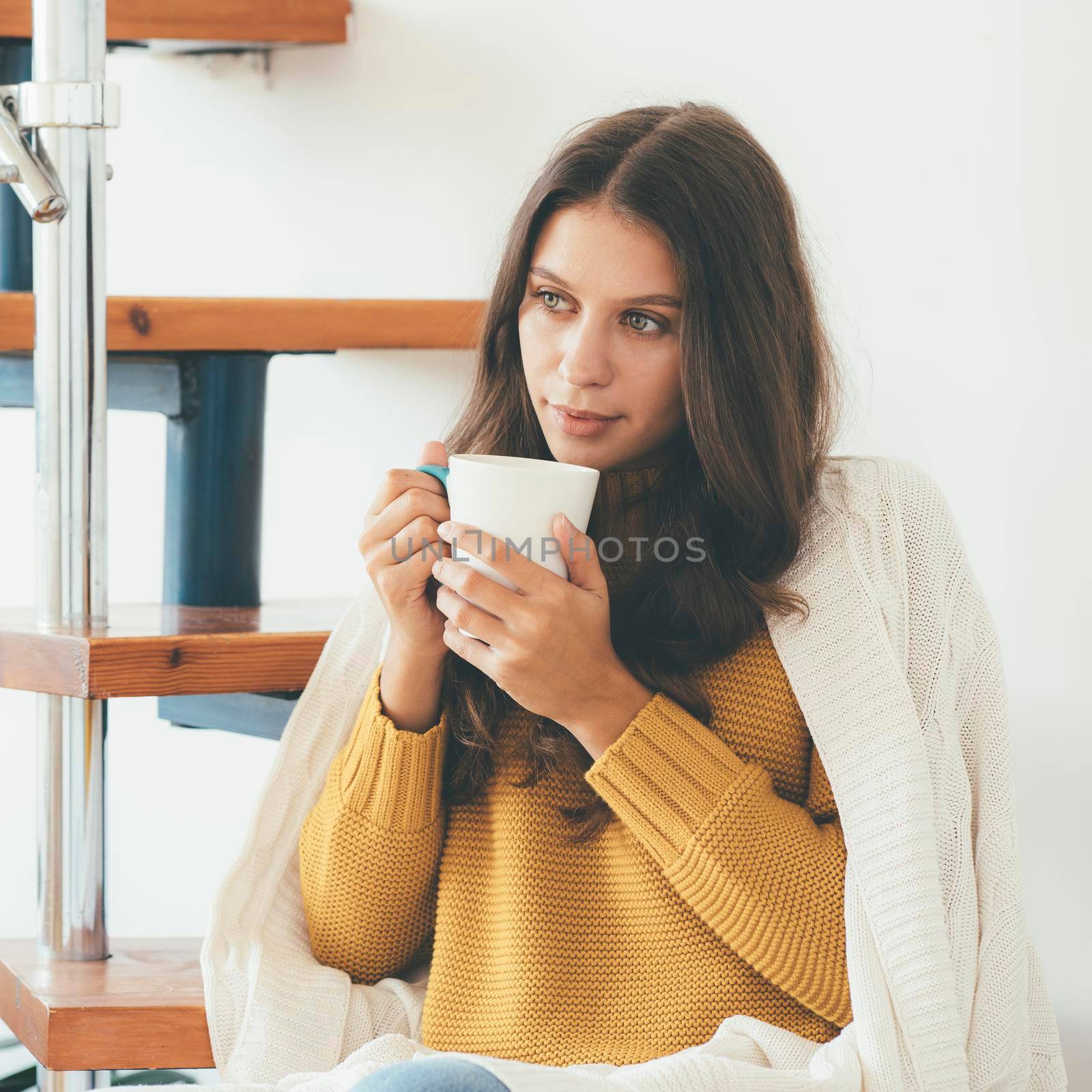 Bautiful young woman sitting onstairs in warm clothes and drinking coffee, wrapped in blanket, warming with hot drink. Weekend at home on cozy autumn or winter day