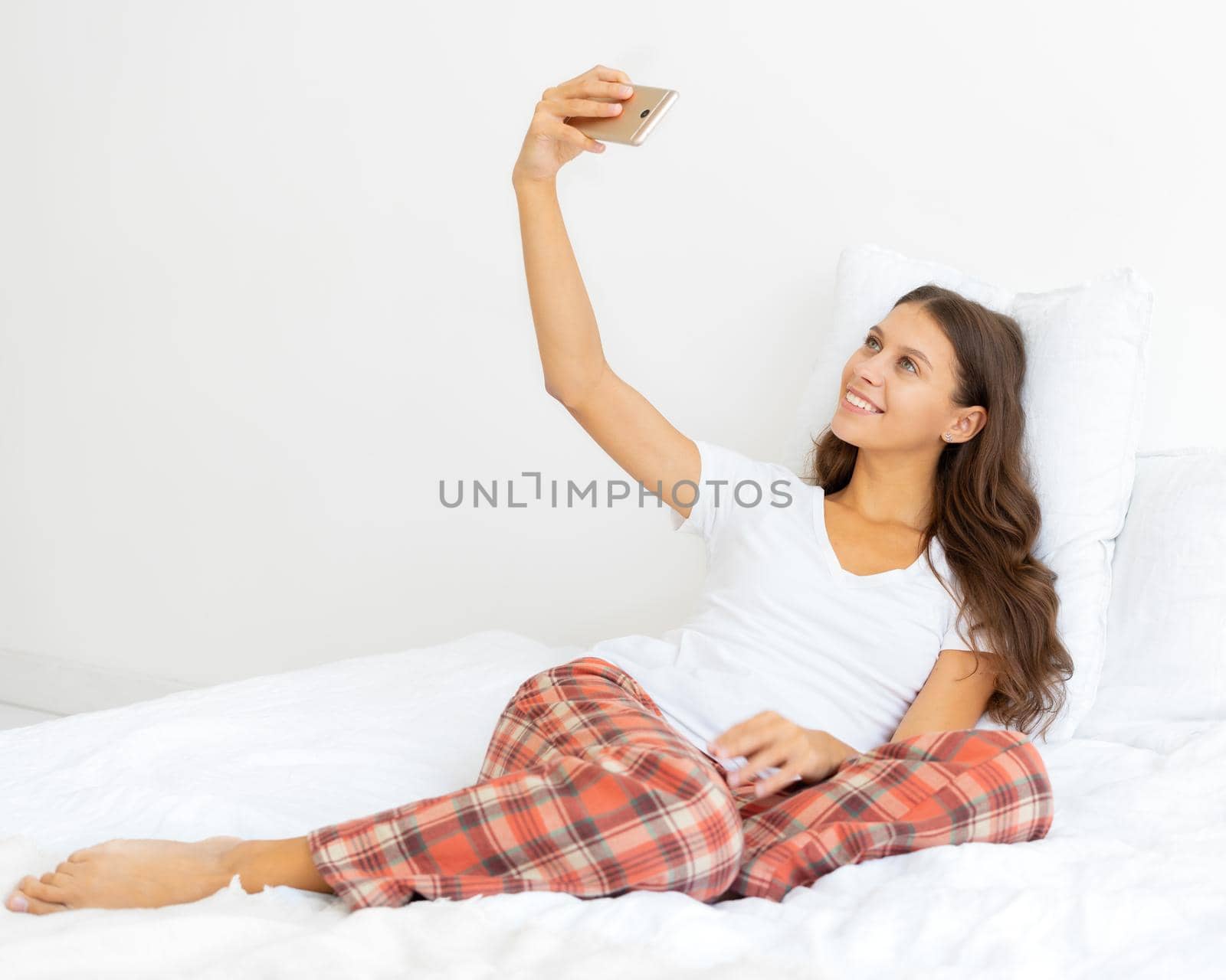 Side view of full lenght pretty female selfie, blonde takes photo on mobile phone laying in bed. Beautiful woman looking at camera and smiling, love for yourself.