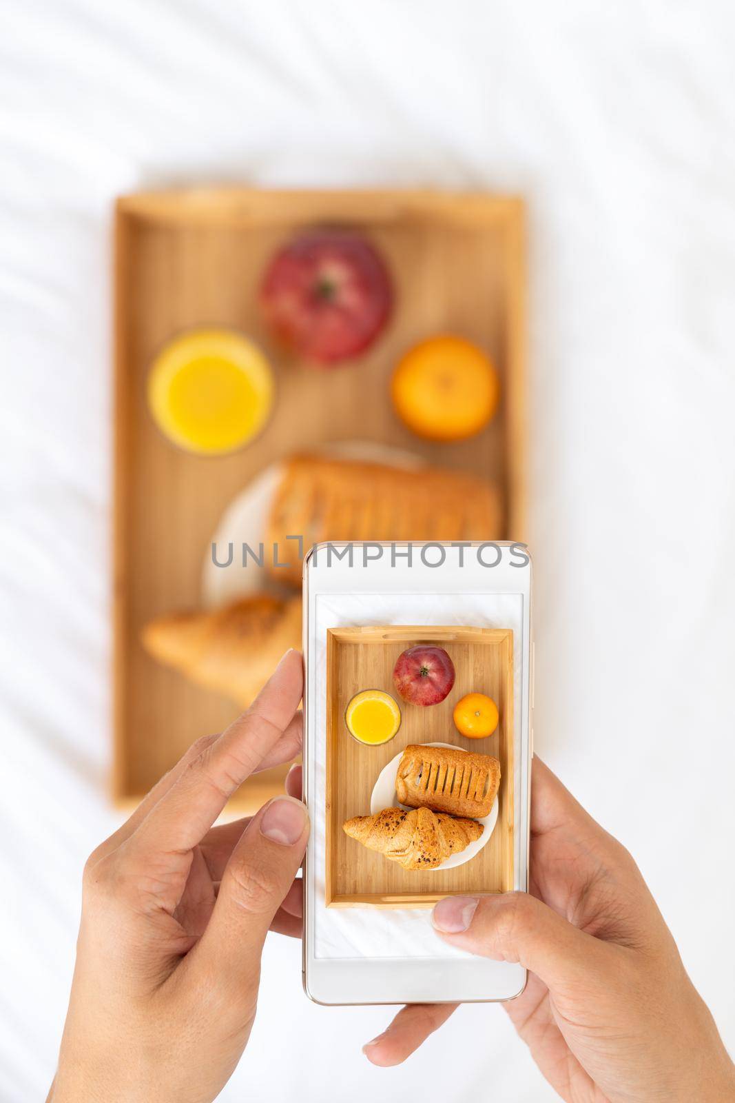 Blogger taking photos of food, shooting Breakfast in bed at hotel on mobile phone, tray with juice, fruit and croissant on white sheets