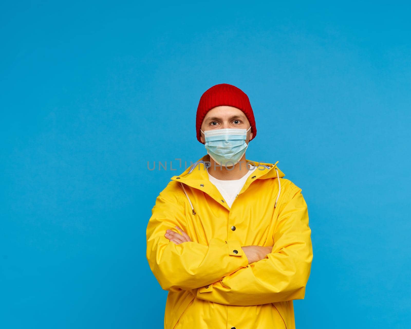 Waisted portrait of man in protective medical mask stands with his arms crossed and looks at camera. Bright blue background, yellow raincoat and red warm hat