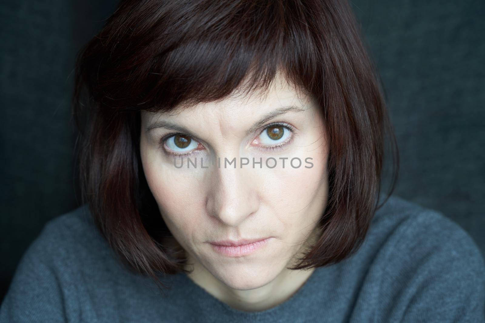 Woman looks from under her brows, frowning and stern. A serious face close up. Anger and wariness. Beautiful caucasian female with dark hair at home during quarantine