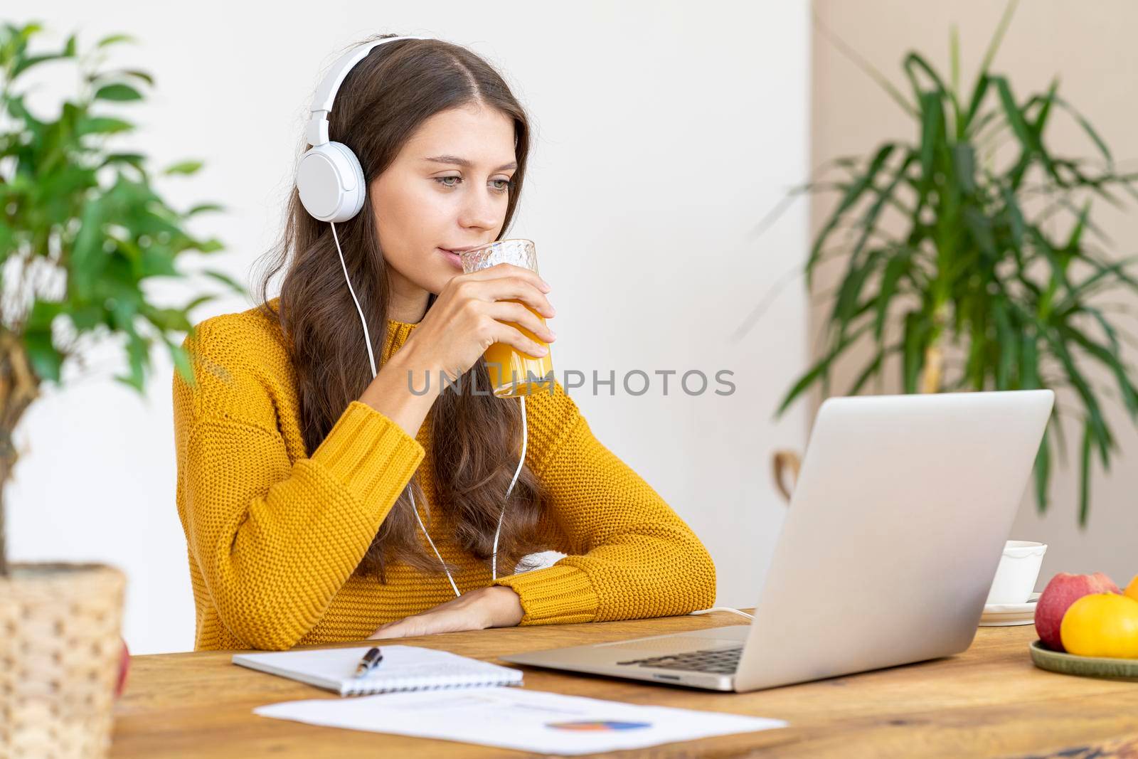 Young girl with headphones talking on conference calls,drinking orange juice, smiling. Beautiful woman with long hair in bright yellow sweater works remotely from home