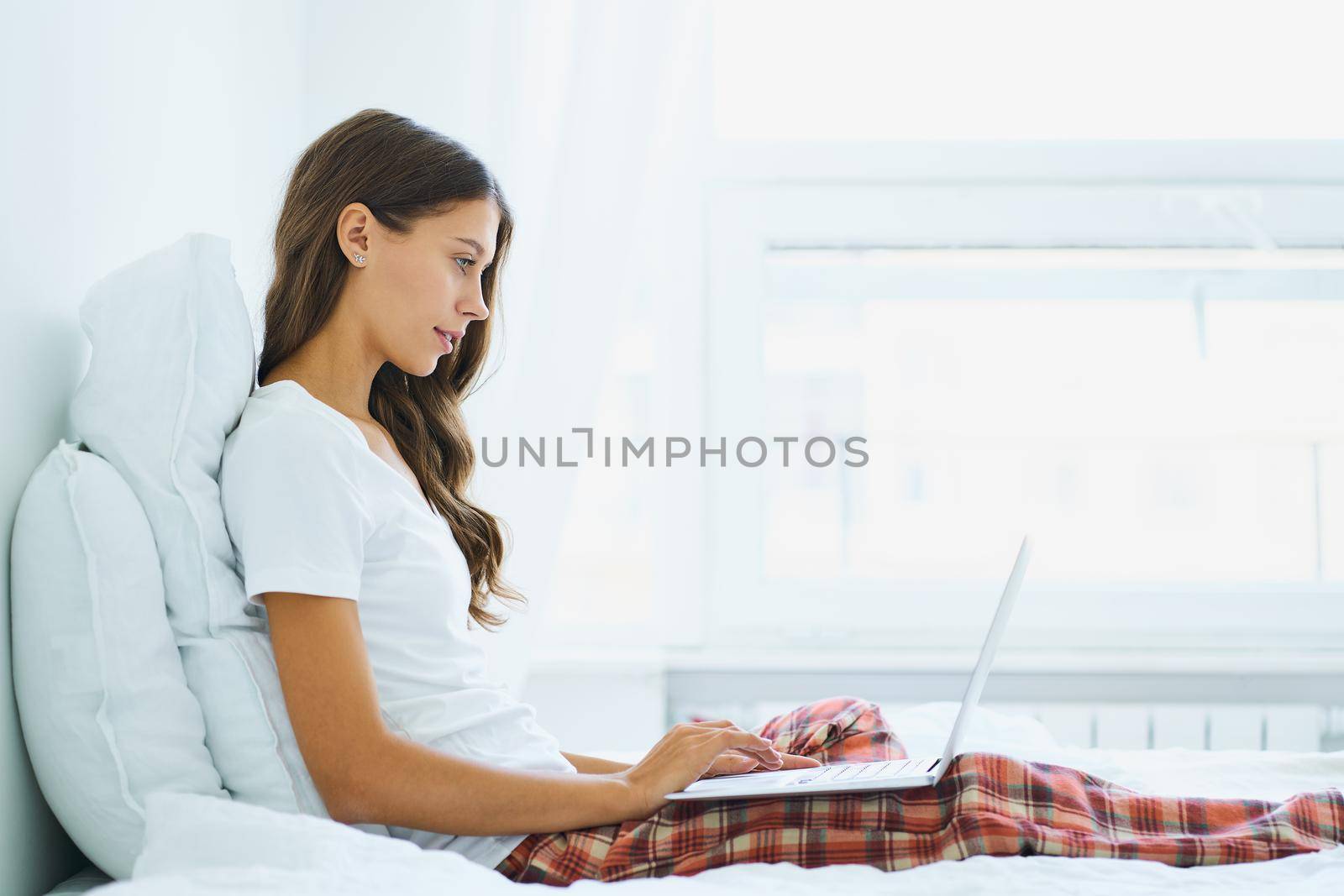 Side view of young woman sitting on bed in bedroom and typing on laptop. Work from home, distance learning for students, surfing Internet in morning after waking up