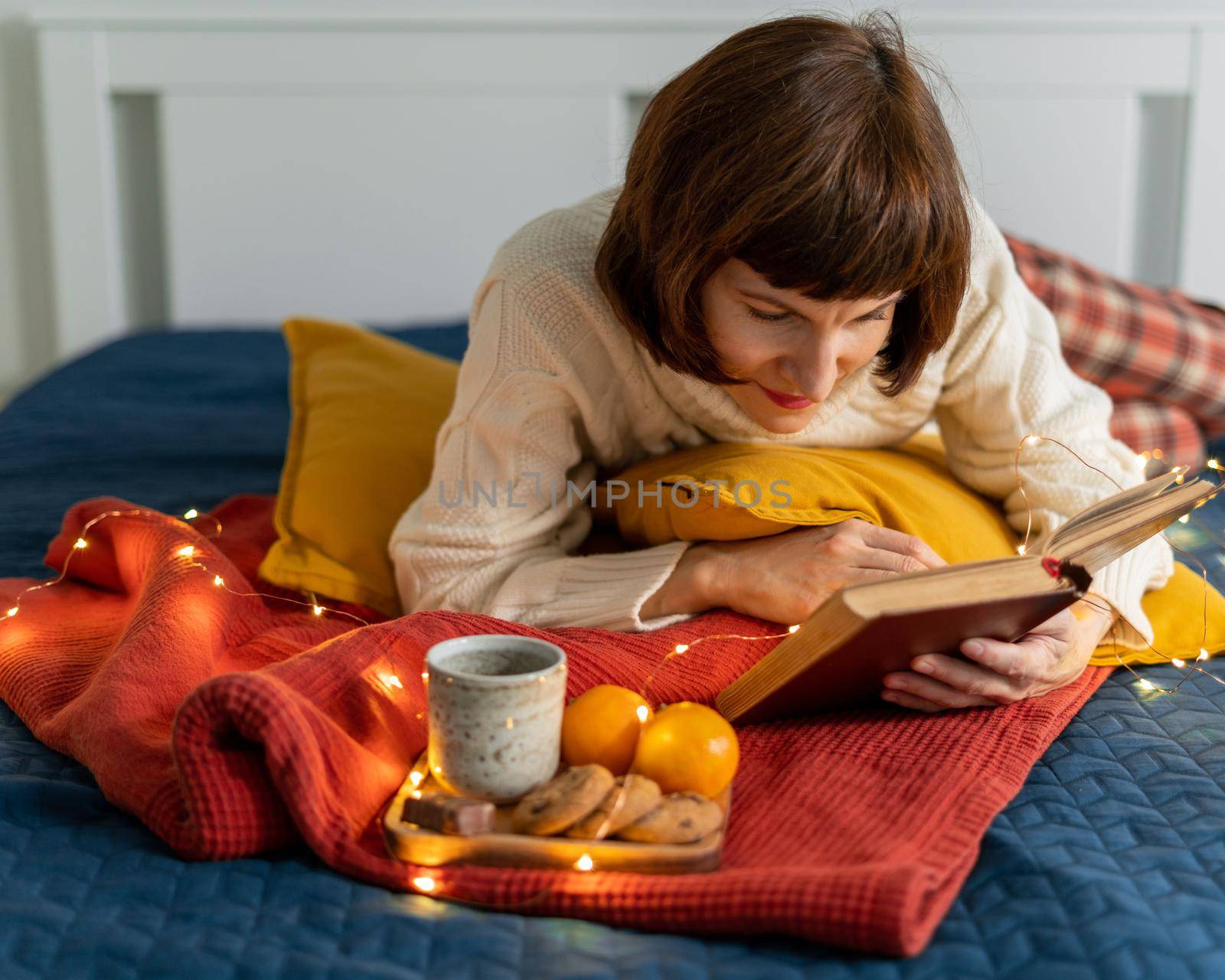 Beautiful mature woman reading book. Cozy background for relaxing and reflection. Stay home concept. Christmas decoration with garland lights, pillows and hot drink with cookies on bed. Dark evening