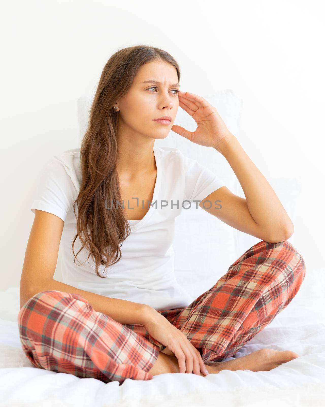 Woman massaging temples of head due to headache after waking up. Migraine. Beautiful young lady in pajamas is sitting on bed and looking away. Vertical