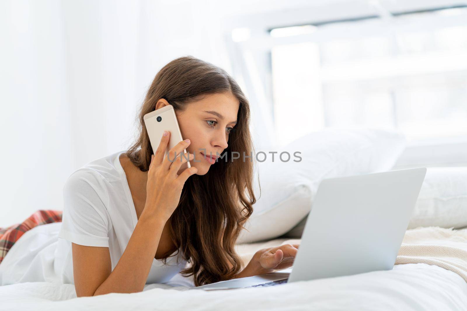 Young woman working at home laying on bed in pajamas, speaking on mobile. Female in bedroom typing on laptop, distance learning for students, surfing Internet in morning after waking up