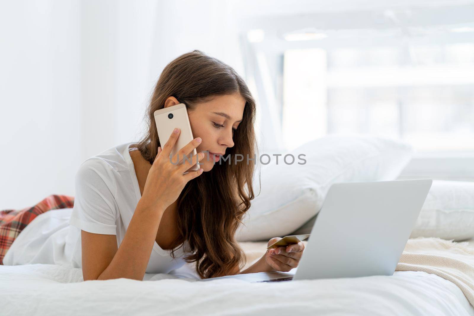 Young woman shopping online, speaking on mobile phone, security call from bank. Female laying on bed in bedroom and working from home on laptop