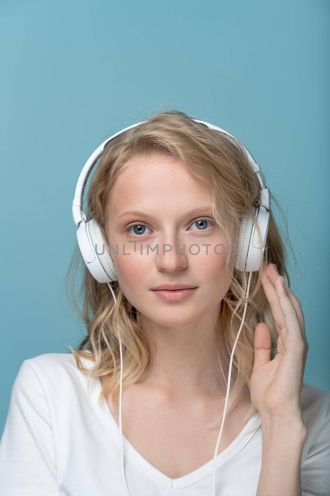 Closeup portrait of young woman closed eyes listening music via headphones on color neutral tone Aqua Menthe wall. Pretty serious clever blonde woman with curly hair looking at camera, vertical