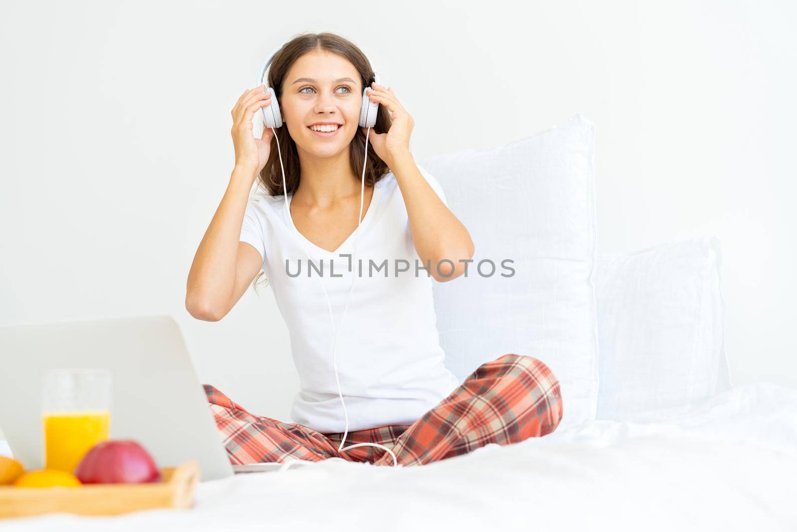 Young smiling woman sitting on bed in bedroom and listening to music. Breakfast in hotel, surfing Internet in morning after waking up