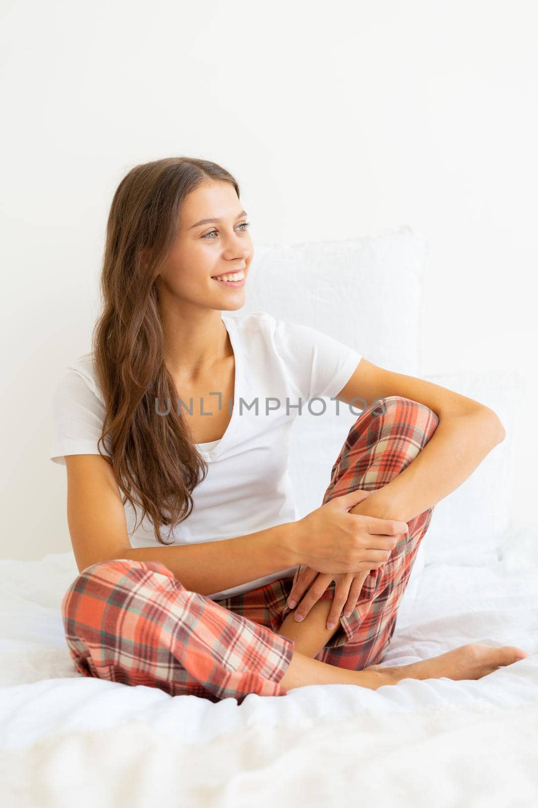 Beautiful young woman without makeup sitting on bed and looking away after waking up. Smiling young female meeting new day in good mood, positive attitude