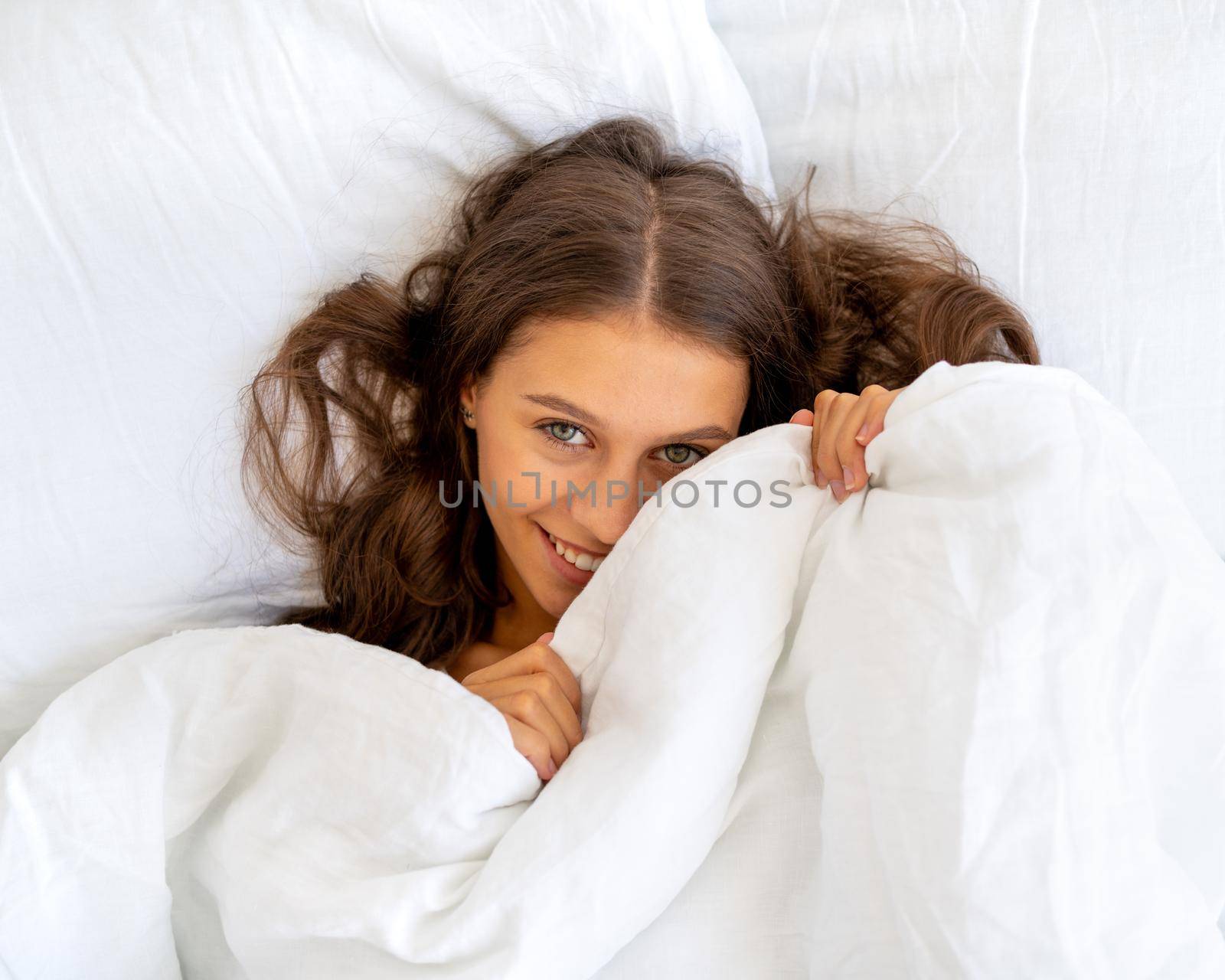 Close up portrait of beautiful smiling woman in bed. Female with long hair resting, good night sleep concept, enjoying fresh soft bedding linen and mattress in bedroom