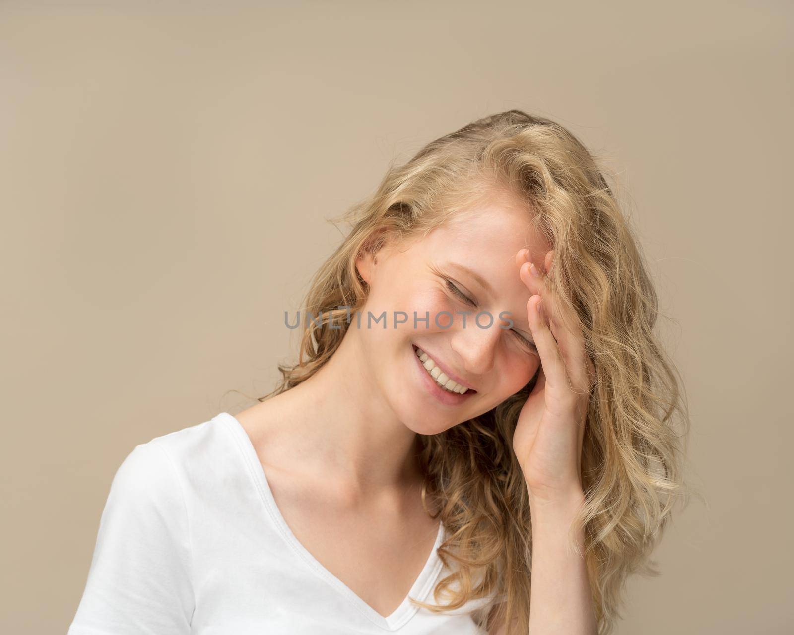 Beautiful young girl laughing. Pretty blonde with curly hair in white t shirt against beige wall smiling and covering face with hand