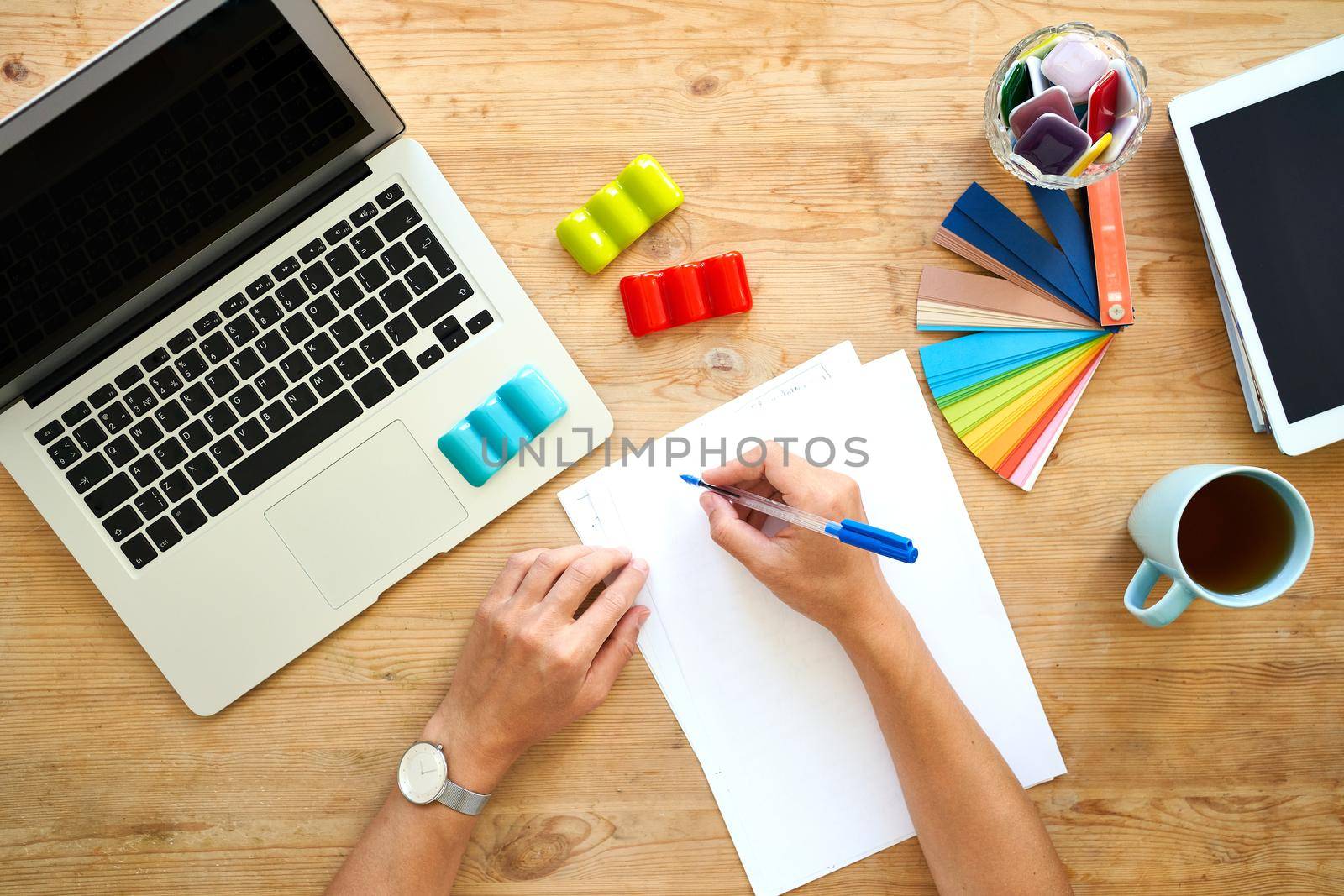 Designer workplace, top view. Home office - desk with laptop, hands with colourful set, color palette selection. Digital tablet and blank white paper for writing