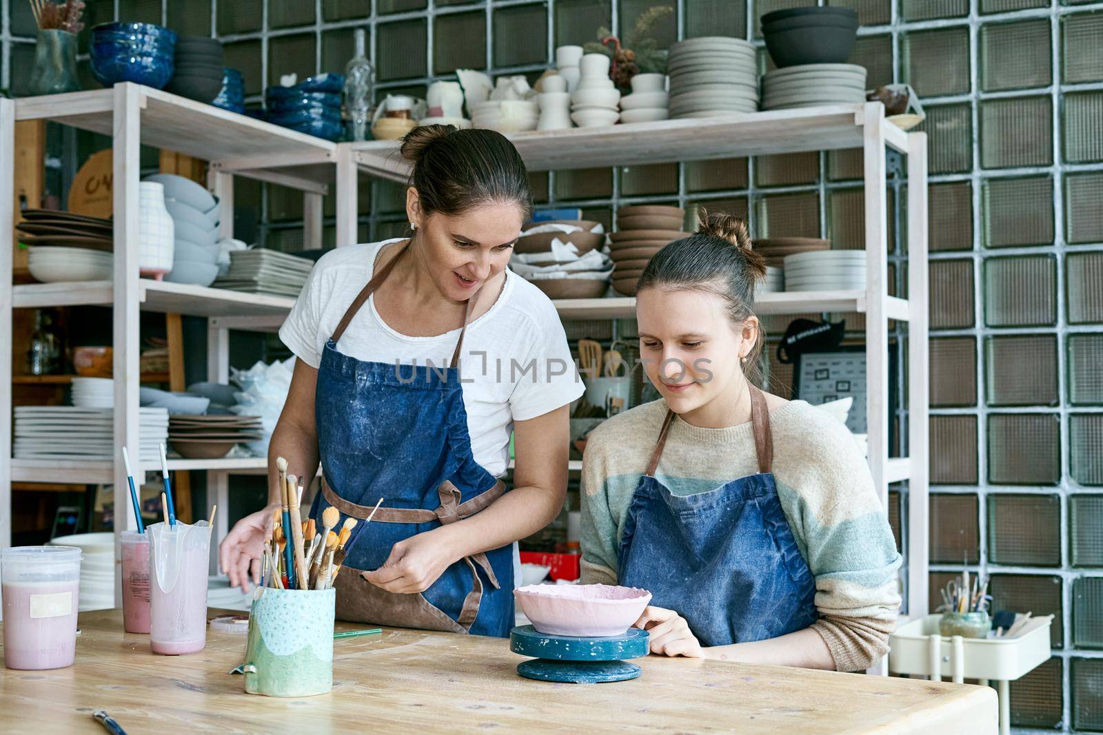 Woman making ceramic pottery. Attractive skilled young lady in apron standing at table and teaching by NataBene