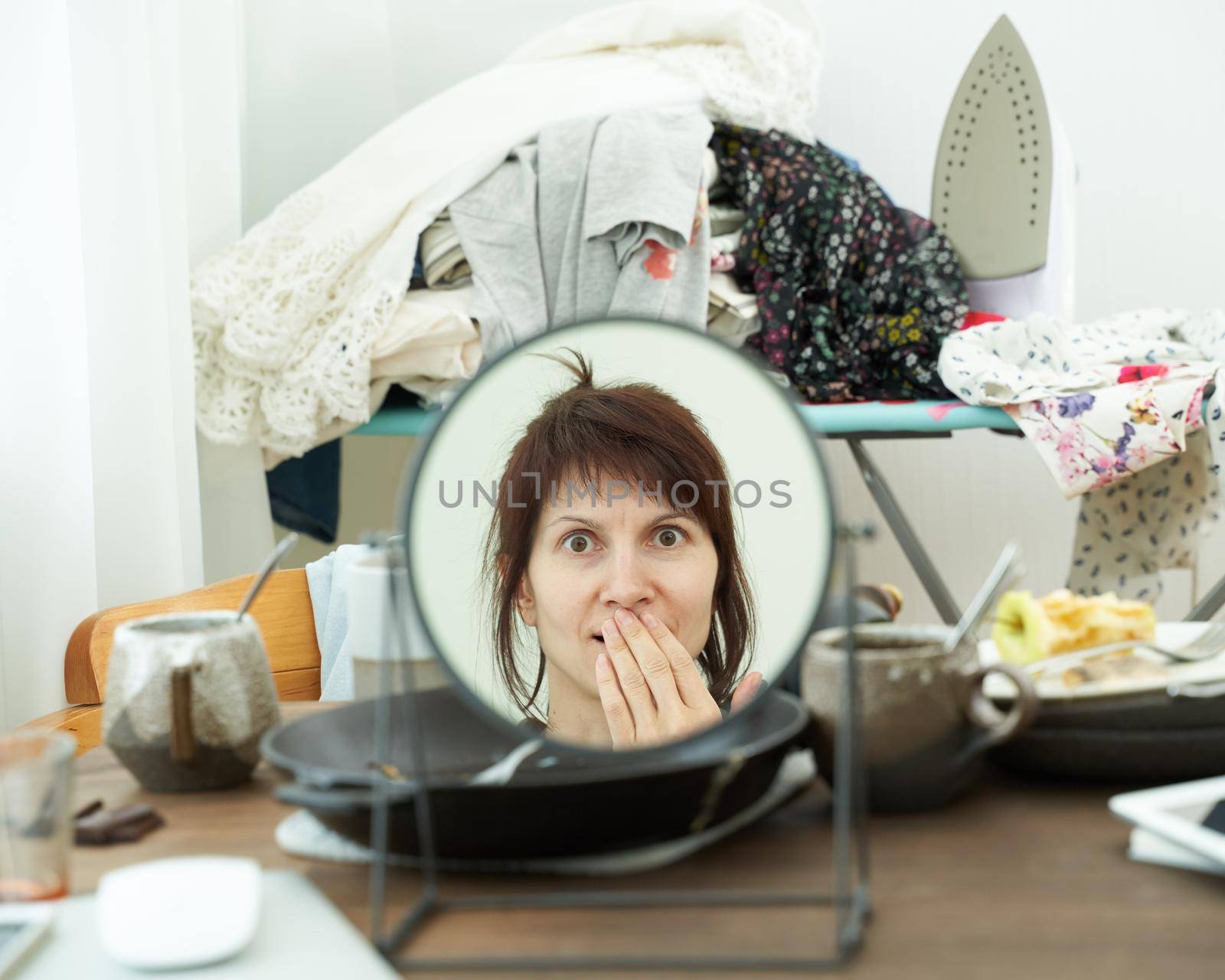Woman looks at herself in mirror against background of mess, dirty dishes, piles of clothes, chaos in room. Concept of abandoned household