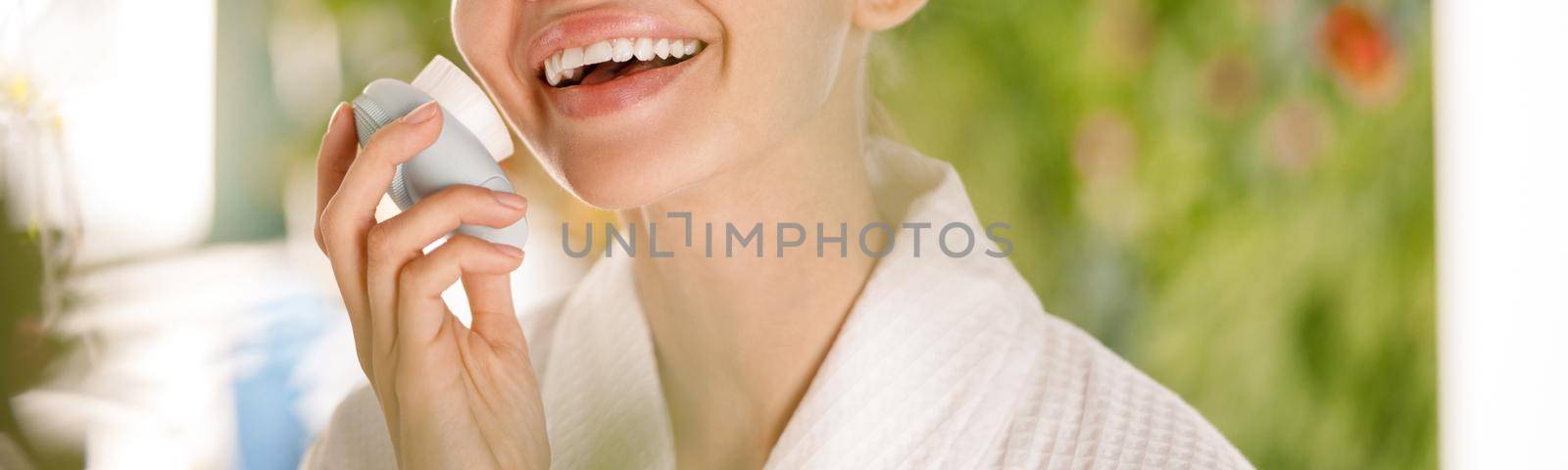 Portrait of lovely young woman with flawless skin wearing bathrobe smiling at camera while using silicone face brush for facial cleansing in the morning. Beauty, skincare, spa concept