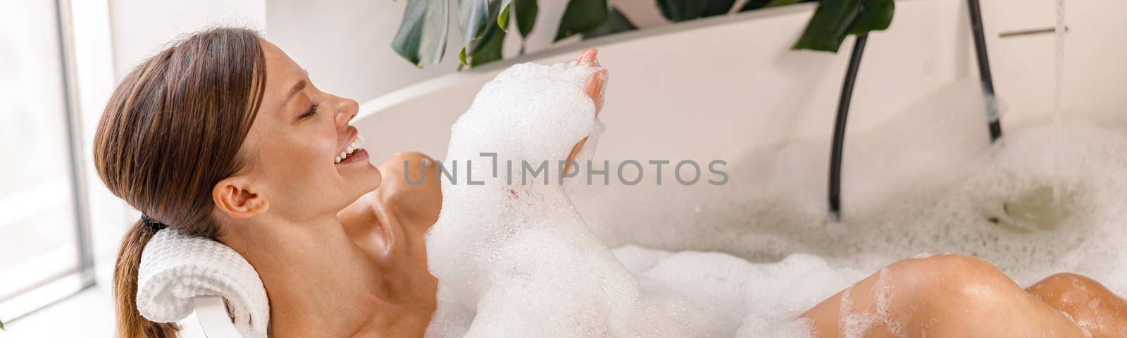 Bathing young woman relaxing in bath, smiling and playing with bubble foam. Body care, wellness concept