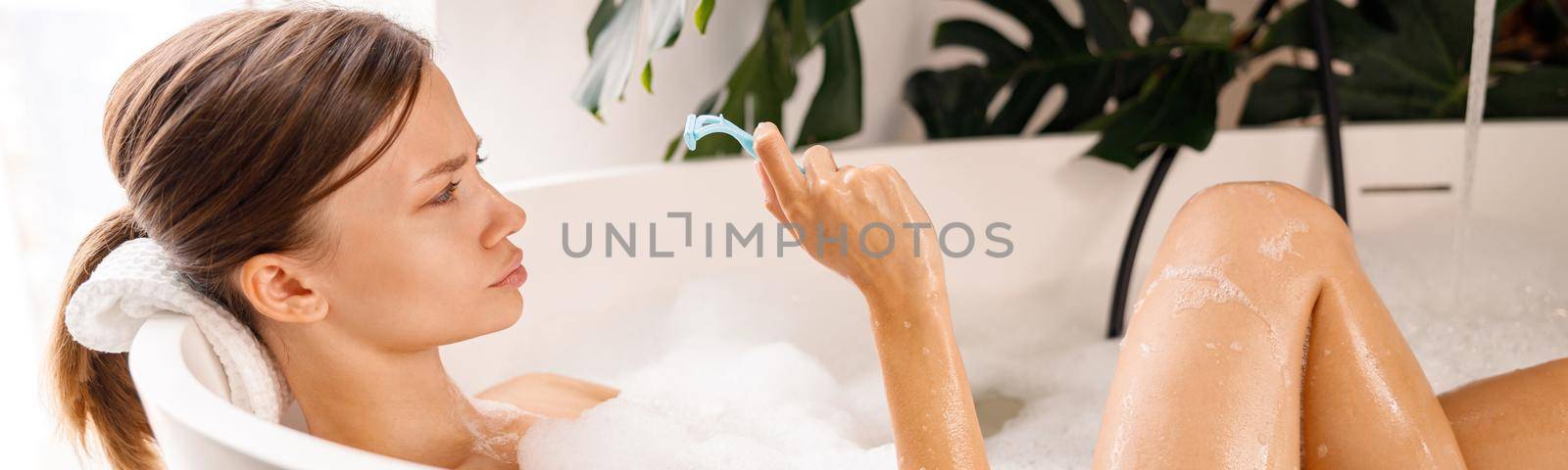 Portrait of young woman making funny faces and holding a razor for shaving body hair while relaxing in bathtub. Spa, beauty concept