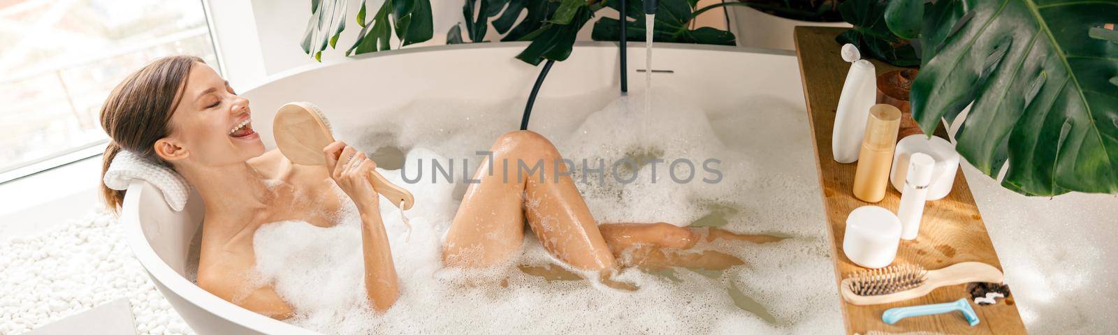 Beautiful young woman having fun, singing while relaxing in bathtub at luxury spa resort. Wellness, body care concept