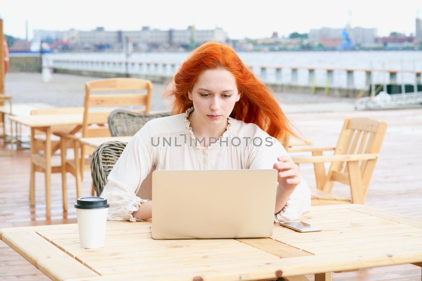 Young readhead woman sitting in cafe outdoors and using laptop while drinking coffee from plastic cup
