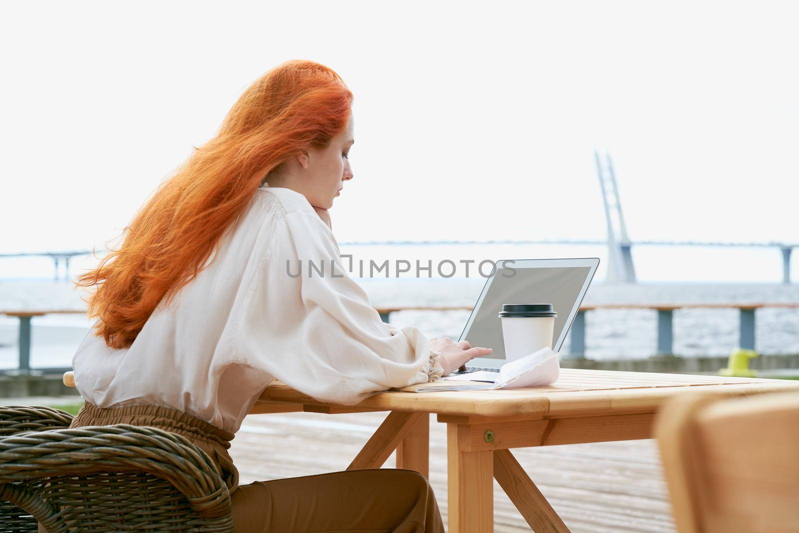 Back view of young redhead female in casual clothes sitting in outside cafe and texting on laptop with plastic cup of coffee